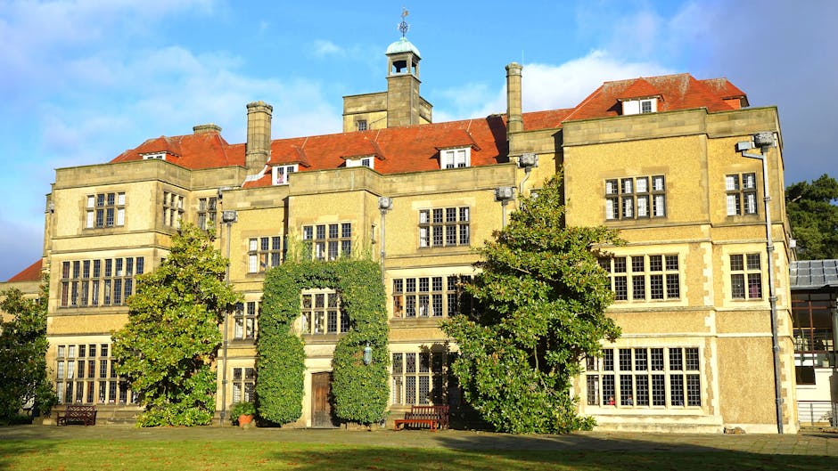 A historic brick mansion surrounded by lush green trees and well-maintained gardens, with large windows and a chimney visible on the roof. The scene is captured during daytime with natural light highl