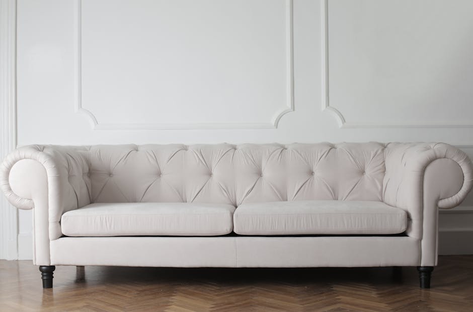A person wearing black gloves and dark jeans is using a handheld electric upholstery cleaner on a dark gray fabric sofa in a residential living room. The sofa has a sleek, modern design with wide armr