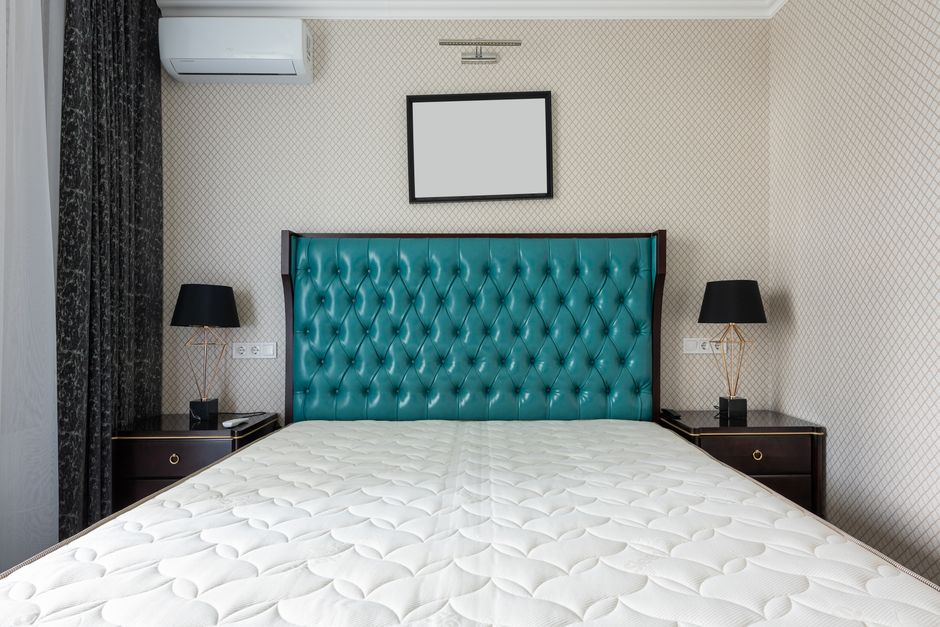 A modern bedroom featuring a clean, white mattress placed directly on the wooden floor. The room has a minimalist design with a purple upholstered headboard, a built-in wooden shelving unit on the rig